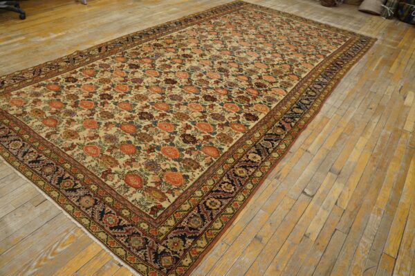 A large low-pile rug featuring a multicolored floral pattern on a cream field and a dark navy border, laid on wooden planks.