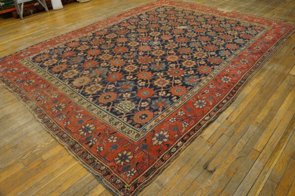 Multicolored floral rug with a dark blue field and wide red border rests on aged yellow wood plank flooring.