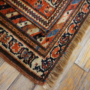 Traditional rug corner featuring rust, navy, and cream geometric patterns with fringe on light wooden flooring.