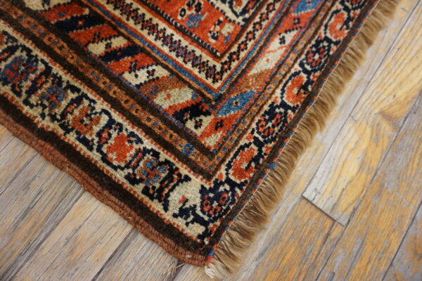Traditional rug corner featuring rust, navy, and cream geometric patterns with fringe on light wooden flooring.