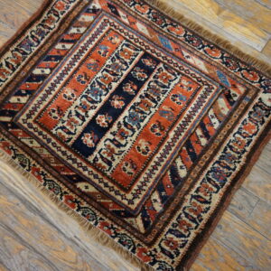 Small square rug featuring orange, navy, and cream geometric patterns rests on distressed wood flooring.