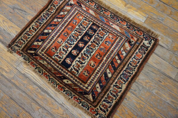 Small square rug featuring orange, navy, and cream geometric patterns rests on distressed wood flooring.