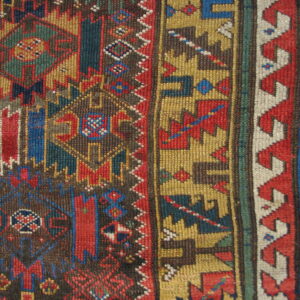 Detail of a short-pile rug with geometric patterns in red, blue, gold, and brown colors on wood floor.