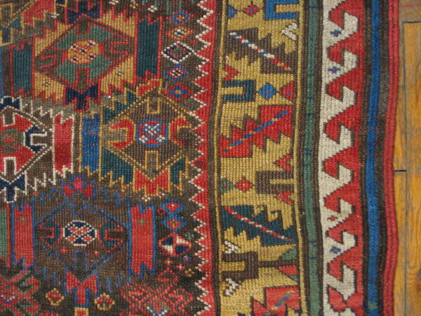 Detail of a short-pile rug with geometric patterns in red, blue, gold, and brown colors on wood floor.