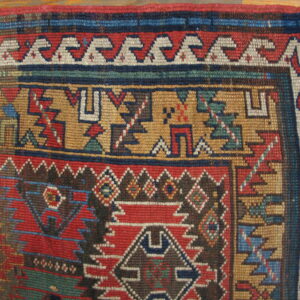 Close-up of a rug corner showing geometric patterns in red, blue, gold, and brown, with visible fringe.