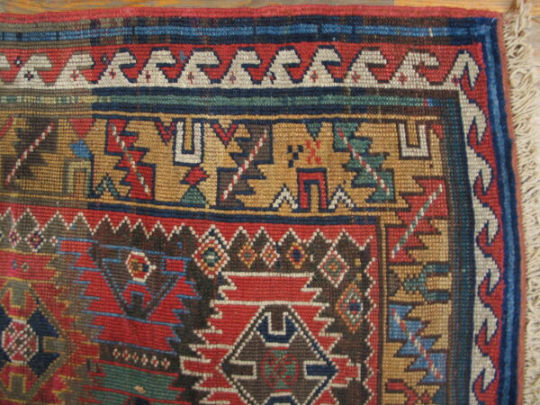 Close-up of a rug corner showing geometric patterns in red, blue, gold, and brown, with visible fringe.