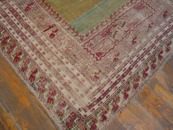 Distressed rug corner featuring magenta and cream geometric borders next to distressed green and tan panels on wood floor.