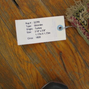 Worn maroon and green patterned rug corner with an attached tag on distressed reddish wood flooring.
