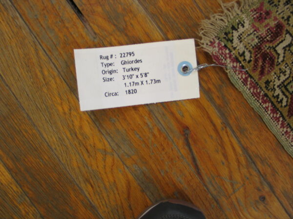 Worn maroon and green patterned rug corner with an attached tag on distressed reddish wood flooring.