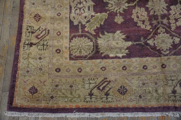 Intricate low-pile rug in tan and burgundy with floral motifs rests on worn wooden floorboards.