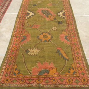 Long olive green runner rug featuring a patterned border and stylized motifs in pink, yellow, and blue on stone flooring.