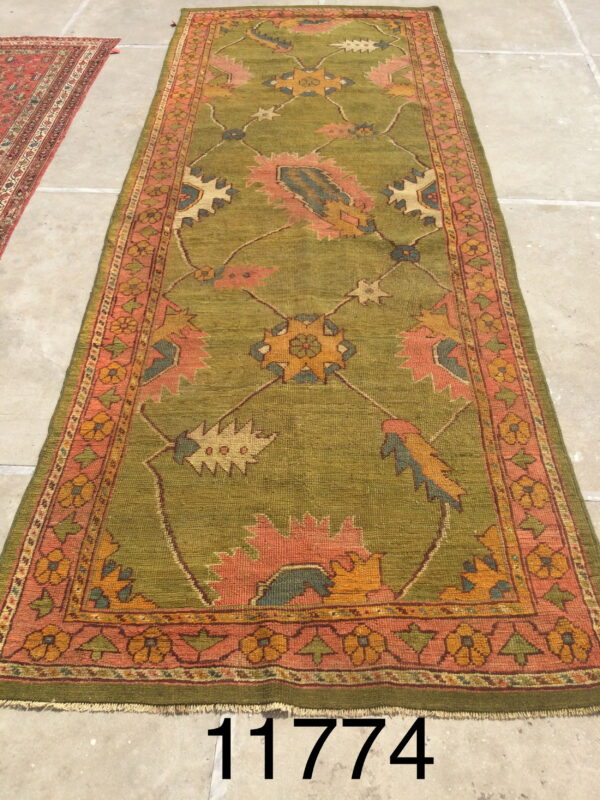 Long olive green runner rug featuring a patterned border and stylized motifs in pink, yellow, and blue on stone flooring.