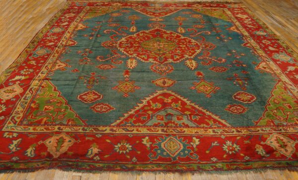 Teal rug with a detailed red, green, and gold medallion and thick red borders on wooden flooring.