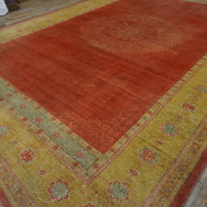 Rust-colored rug with a faded central pattern and a wide gold-green geometric border on wood flooring.