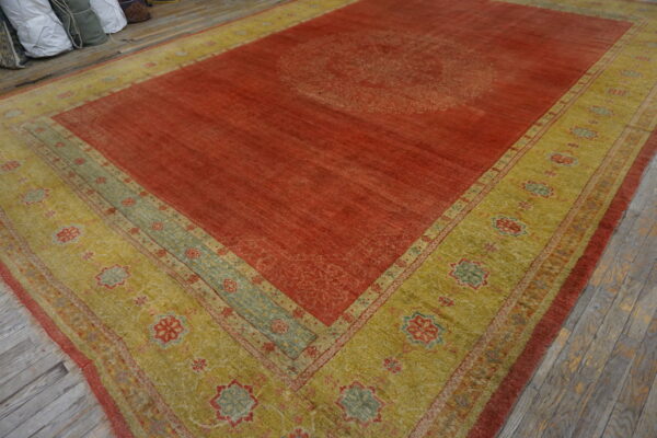 Rust-colored rug with a faded central pattern and a wide gold-green geometric border on wood flooring.