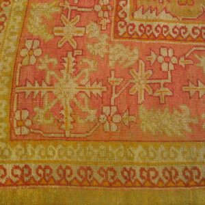 Tightly woven rug corner featuring pink, yellow, and cream geometric and floral patterns on wood flooring.