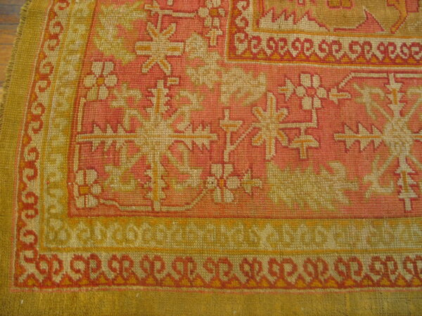 Tightly woven rug corner featuring pink, yellow, and cream geometric and floral patterns on wood flooring.