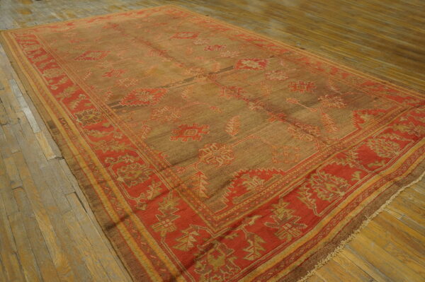 Brown-tan rug with red and gold geometric patterns and a wide border rests on worn wooden floorboards.