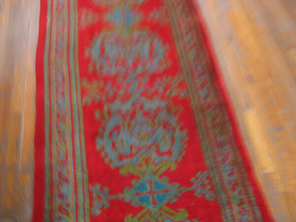 A blurry red runner rug with abstract blue and green traditional patterns lies on a light wood floor.