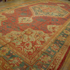 Large red, teal, and tan patterned rug lying on worn light brown hardwood flooring.