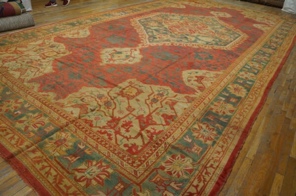Large red, teal, and tan patterned rug lying on worn light brown hardwood flooring.