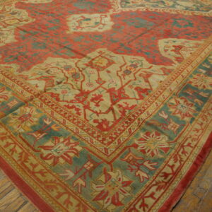 Ornate traditional rug with brick red field, teal border, and tan motifs, lying on wood plank flooring.