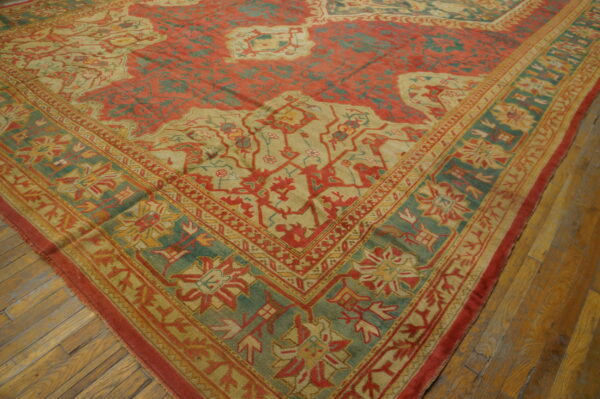 Ornate traditional rug with brick red field, teal border, and tan motifs, lying on wood plank flooring.