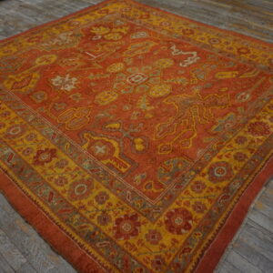 Ornate square rug in rust and golden yellow with geometric patterns rests on distressed gray wood.