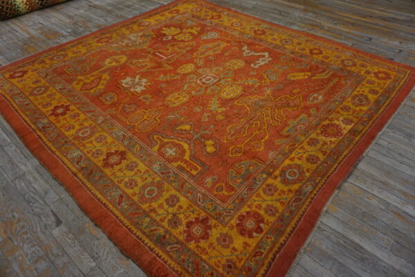 Ornate square rug in rust and golden yellow with geometric patterns rests on distressed gray wood.