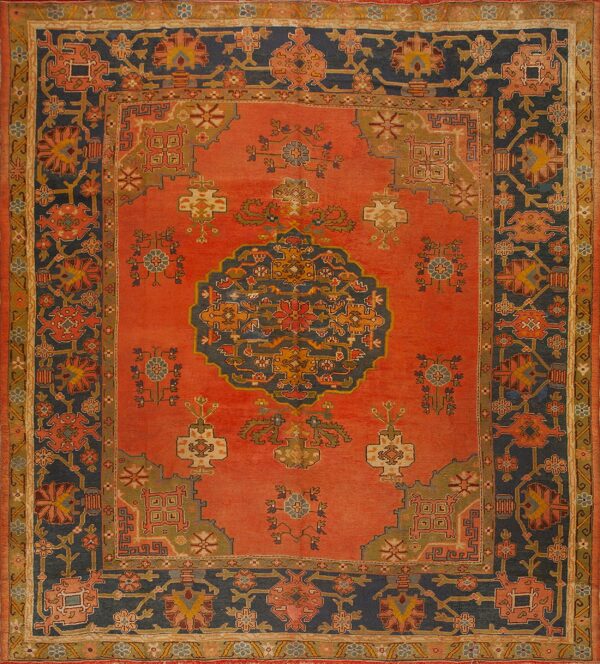 Ornate traditional rug with a coral field, stylized floral motifs, and wide indigo borders.
