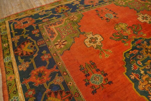 Rust orange patterned rug with stepped navy and moss green borders and geometric motifs on a wood floor.