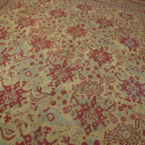 Red and blue geometric rug with a tan field and elaborate borders sits on brown hardwood floors.