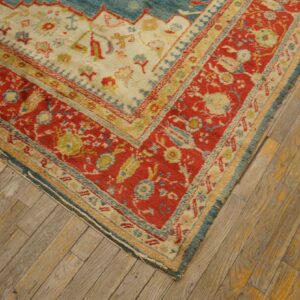 The corner of a low pile rug featuring red, cream, and blue patterns rests on distressed wooden planks.