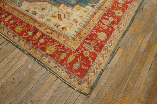 The corner of a low pile rug featuring red, cream, and blue patterns rests on distressed wooden planks.