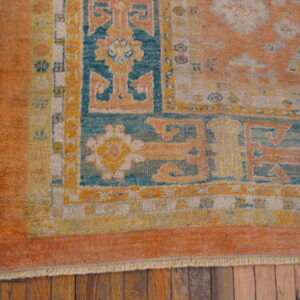 Low-pile rug corner showing geometric borders in orange, teal, and yellow on rustic brown wood flooring.