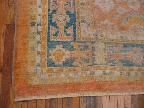 Low-pile rug corner showing geometric borders in orange, teal, and yellow on rustic brown wood flooring.