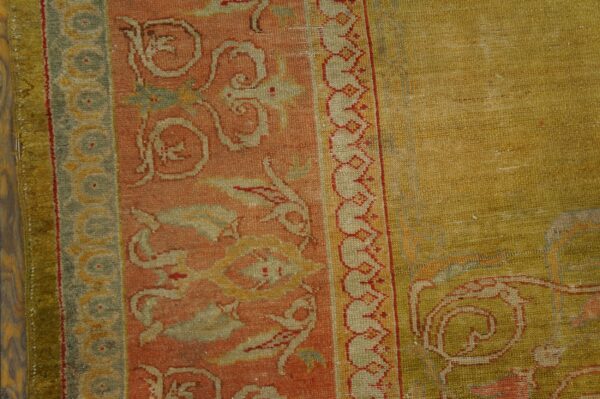 Muted rust and sage green patterned borders meet a textured, olive gold central field on wood flooring.