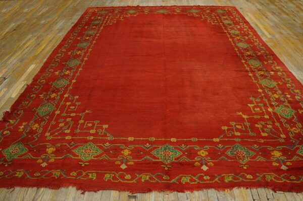 Low-pile red rug with green and yellow geometric borders on worn wooden planks.