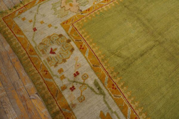 Textured green rug with patterned borders in cream, ochre, and red, resting on wooden flooring.