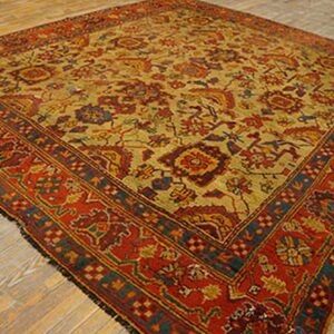 Square low-pile rug in mustard yellow and deep red with intricate traditional blue and orange patterns on wood floor.
