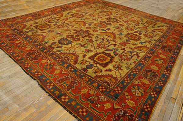Square low-pile rug in mustard yellow and deep red with intricate traditional blue and orange patterns on wood floor.
