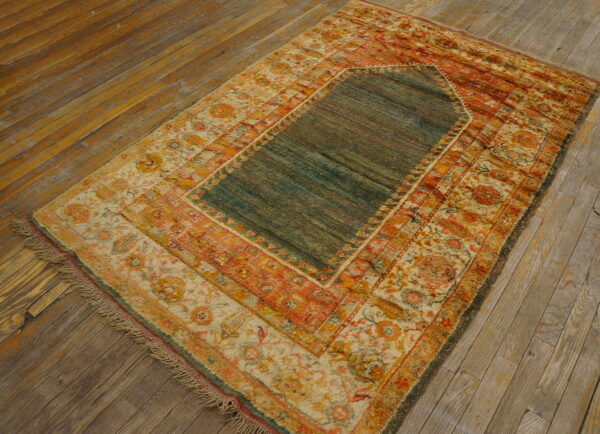 Prayer-style rug in teal, terracotta, and gold with ornate borders rests on distressed wooden flooring.