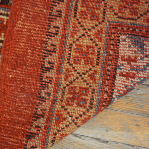 A corner of a textured red and tan geometric rug rests on light distressed wood planks.