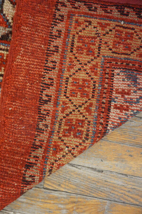 A corner of a textured red and tan geometric rug rests on light distressed wood planks.