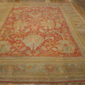 Low-pile area rug in muted red, tan, and sage green, featuring an intricate overall floral design on wooden flooring.