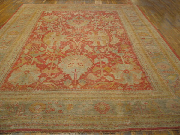 Low-pile area rug in muted red, tan, and sage green, featuring an intricate overall floral design on wooden flooring.