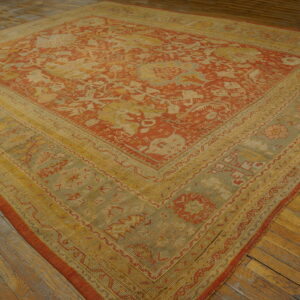 Square rug with rust, yellow, and green traditional patterns rests on aged, wide-plank hardwood floors.