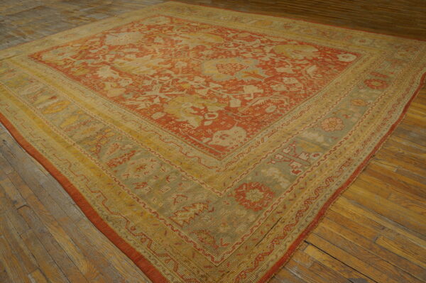 Square rug with rust, yellow, and green traditional patterns rests on aged, wide-plank hardwood floors.