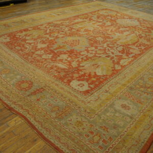 Ornate square rug with a terracotta red field and complex yellow-green borders on wood planks.