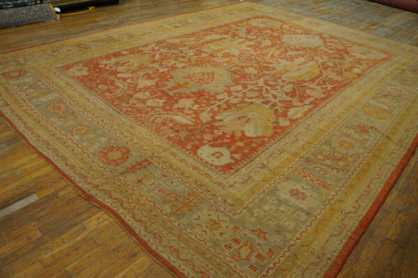 Ornate square rug with a terracotta red field and complex yellow-green borders on wood planks.